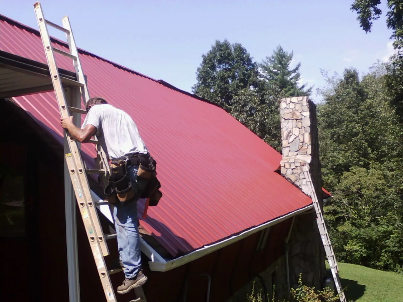 Roofer on a ladder beside a red metal roof and stone chimney — Soffit & Fascia Repair by Roofing Brothers in Raymondville